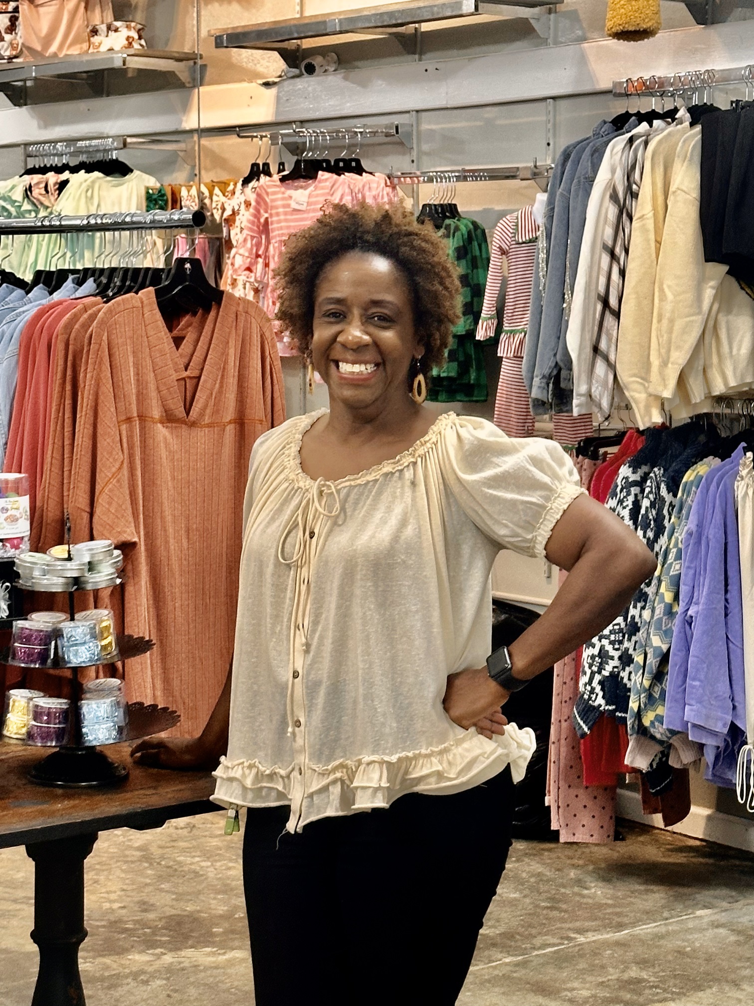 A woman stands smiling inside Collective & Vine boutique, wearing a cream-colored, ruffled blouse and black pants. Behind her are racks filled with colorful women’s clothing including dresses, sweaters, and tops. A display table beside her showcases small tins of candles or balms. The store's interior has a cozy, curated feel with a mix of textures and hues.