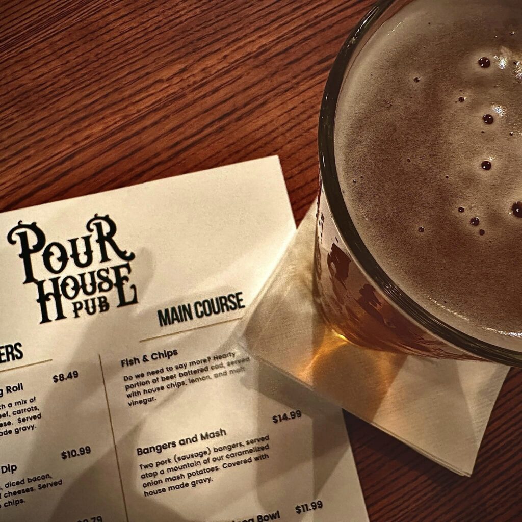 A pint of beer sits on a napkin beside a wooden table at Pour House Pub in Statesboro, partially covering a printed menu featuring dishes like Fish & Chips and Bangers and Mash.
