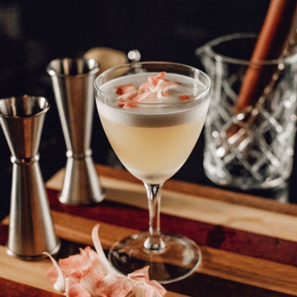 A frothy gin cocktail garnished with flower petals is served in a coupe glass. The drink sits on a striped wooden board surrounded by metal jiggers and a mixing glass with a bar spoon, creating an elegant bar setup at Tandoor & Tap Indian Restaurant.