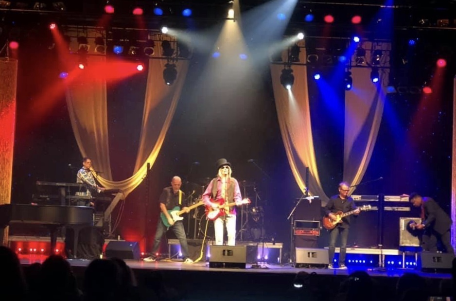 A lively performance during the 'Rockin’ Out Alzheimer’s' event, featuring a band of five musicians on a brightly lit stage. The lead performer, wearing a top hat and a pink jacket, sings and plays guitar at the center. The stage is adorned with flowing golden drapes, with vibrant red, blue, and white spotlights adding energy to the scene. Other band members include a keyboardist, two guitarists, and a member adjusting equipment, creating an engaging atmosphere for a meaningful cause.