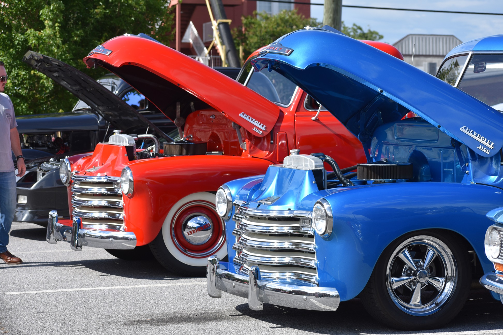 A vibrant photo from BoroFest 2024: Bringing Life to Downtown Statesboro, showcasing a lineup of classic Chevrolet trucks with their hoods open. A bright red truck with whitewall tires and a shiny blue truck with chrome rims stand out under the sunlight. In the background, other classic vehicles and a small crowd of spectators add to the lively atmosphere of the event, surrounded by trees and downtown buildings.