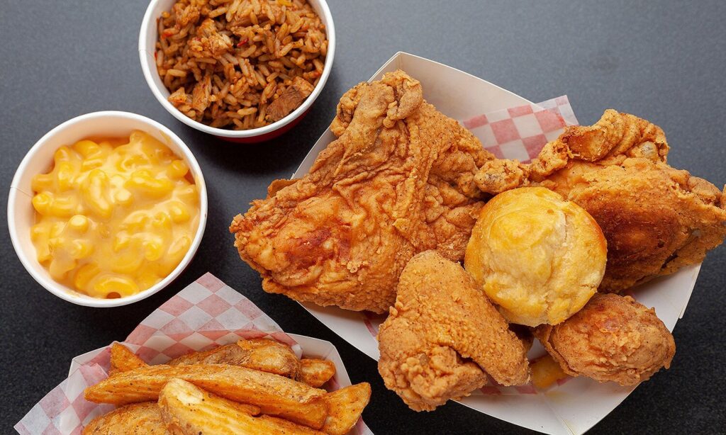 A hearty meal from Krispy Krunchy Chicken featuring a tray of golden fried chicken pieces served with a fluffy biscuit. The meal is accompanied by generous sides, including creamy mac and cheese, seasoned potato wedges, and a cup of flavorful Cajun-style rice. The presentation highlights the crispy texture and rich, comforting flavors, making it a classic Southern-inspired feast.