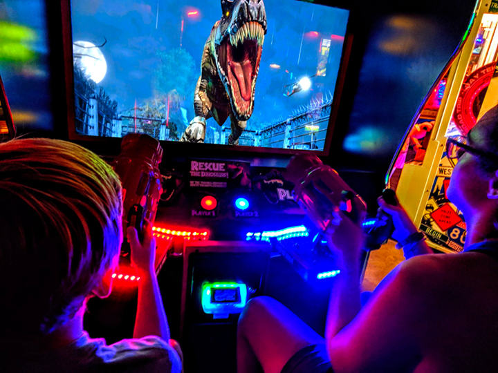 Two kids fully immersed in an arcade game at a venue in The Boro. They are holding brightly lit toy guns, aiming at a screen where a massive, roaring T-Rex charges toward them in a vividly detailed jungle setting. The game, titled 'Rescue the Dinosaurs,' is surrounded by glowing red and blue neon lights, creating an exciting and action-packed environment. The vibrant atmosphere reflects the fun and engaging activities available for kids in the area.