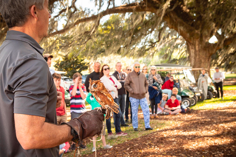 A lively outdoor event in Statesboro where a group of adults and children gather to watch a wildlife demonstration under a canopy of moss-draped oak trees. The foreground shows a handler wearing a gray shirt and leather glove holding a small, reddish-brown owl. The audience, some seated and others standing, looks on with fascination and curiosity. Golf carts and colorful attire add to the casual and family-friendly atmosphere, highlighting the community's engaging and educational activities.