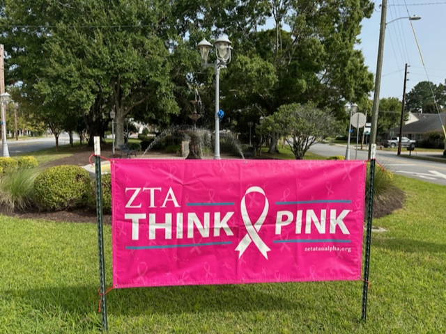 A bright pink banner displayed on a grassy lawn, promoting Zeta Tau Alpha's 'Think Pink' campaign. The banner features a large pink ribbon symbol, the organization's name (ZTA), and the slogan 'Think Pink,' with a website link (zetataualpha.org) in the bottom corner. In the background, a small fountain, trees, and a suburban street add to the peaceful outdoor setting.