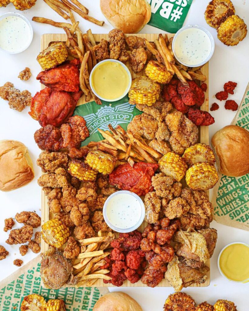 A vibrant platter of Wingstop's signature offerings, featuring an assortment of crispy fried chicken wings in various flavors, golden corn on the cob, seasoned fries, and dipping sauces. The board is surrounded by buns and extra sides, showcasing a delicious and indulgent feast. The bright presentation is enhanced with Wingstop-branded paper, adding a touch of branding to the appetizing spread.