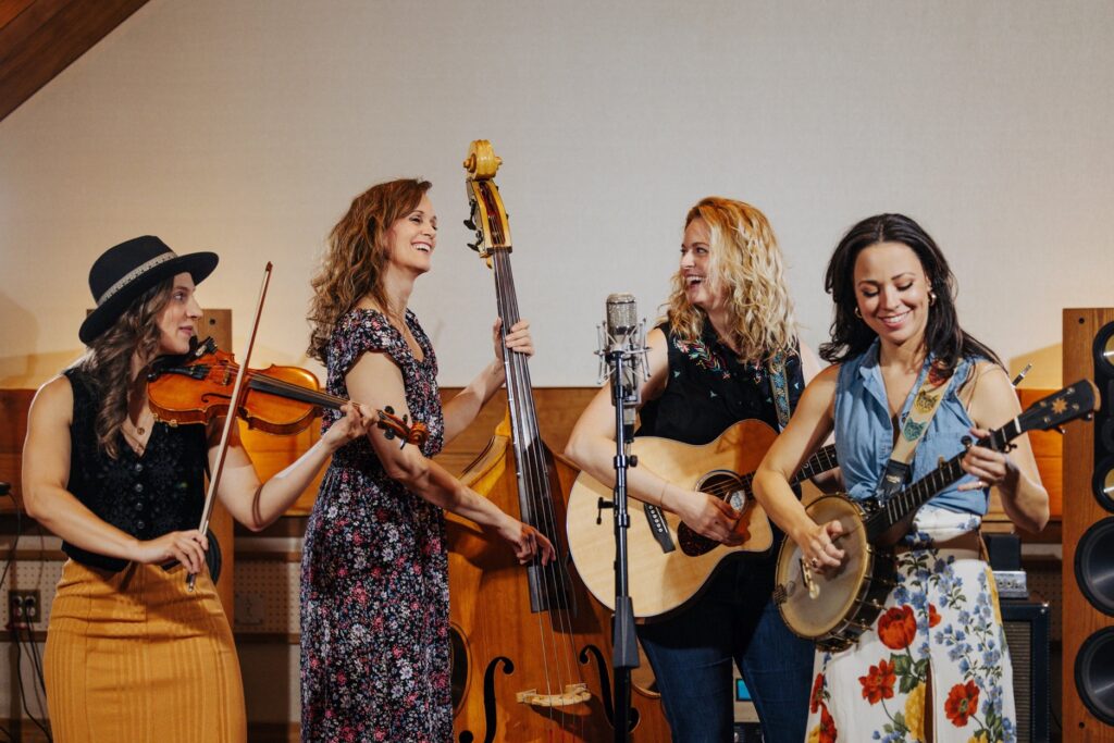 A lively quartet of female musicians performing in an intimate setting, showcasing their talent with string instruments. One plays the violin, another the upright bass, the third strums an acoustic guitar, and the fourth plucks a banjo. Their attire is vibrant and casual, with floral patterns and a mix of modern and vintage styles. A microphone stands at the center, emphasizing the group's harmonized performance as part of Georgia Southern's Performing Arts Center 2024 season.