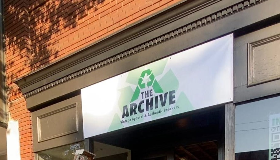 The storefront of 'The Archive,' a new shop in Downtown Statesboro, showcasing a banner with the store's logo. The banner features a green recycling symbol and bold black text reading 'THE ARCHIVE,' along with the tagline 'Vintage Apparel & Authentic Sneakers.' The building facade includes classic brickwork and detailed trim, adding a vintage charm to the shop's modern and eco-friendly branding.