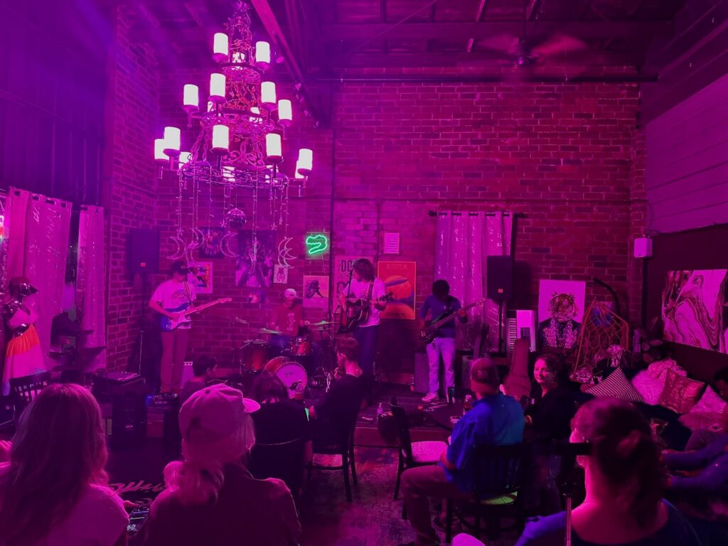 An intimate live music performance at Gators & Gypsies, featuring a five-piece band performing under vibrant purple lighting. The venue has a cozy ambiance with exposed brick walls adorned with eclectic artwork and neon signs. A large ornate chandelier adds a touch of elegance, while an attentive audience sits on mismatched chairs, enjoying the performance.