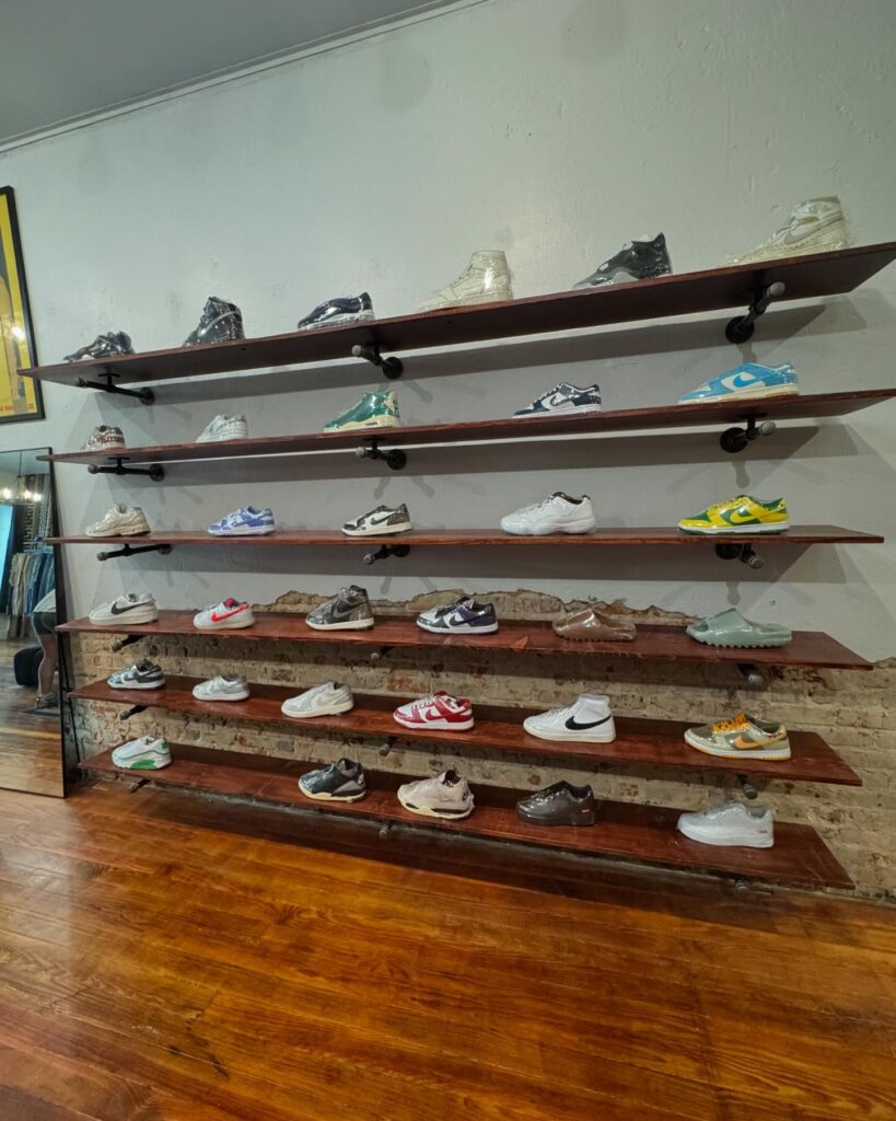 Display shelves in Archive Boro showcasing a collection of sneakers. Wooden shelves are mounted on a textured brick wall, presenting stylish and colorful shoes, including various Nike and trendy designs. The polished wooden floor adds a warm contrast to the sleek sneaker arrangement.