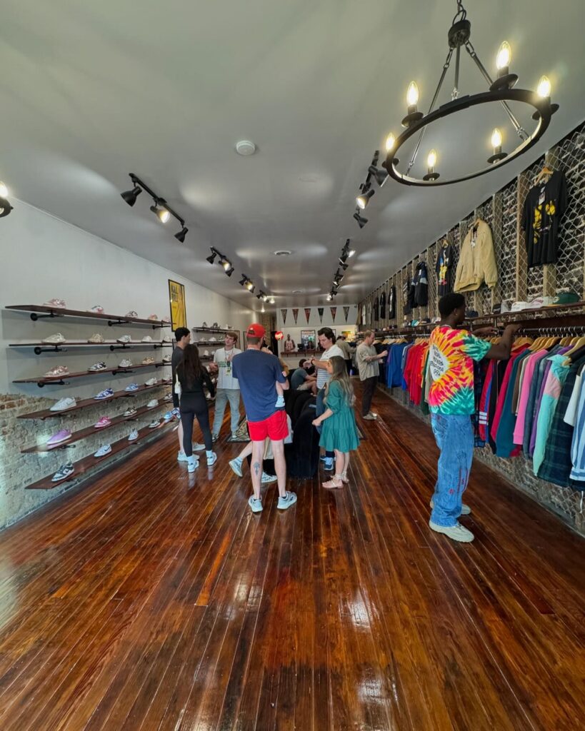 Interior view of Archive Boro, a sleek and modern vintage shop with polished wooden floors. Shelves on the left display a variety of sneakers, while racks on the right showcase colorful jackets and apparel. A small group of customers browse the merchandise, adding a lively atmosphere under stylish track and chandelier lighting.