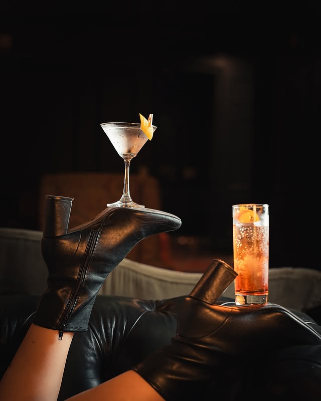 A creative setup at Oak 45 featuring two stylish drinks balanced on a pair of black high-heeled boots. A martini garnished with a lemon twist sits on one heel, while a tall, vibrant cocktail with orange slices rests on the other, all against a dimly lit, sophisticated background.