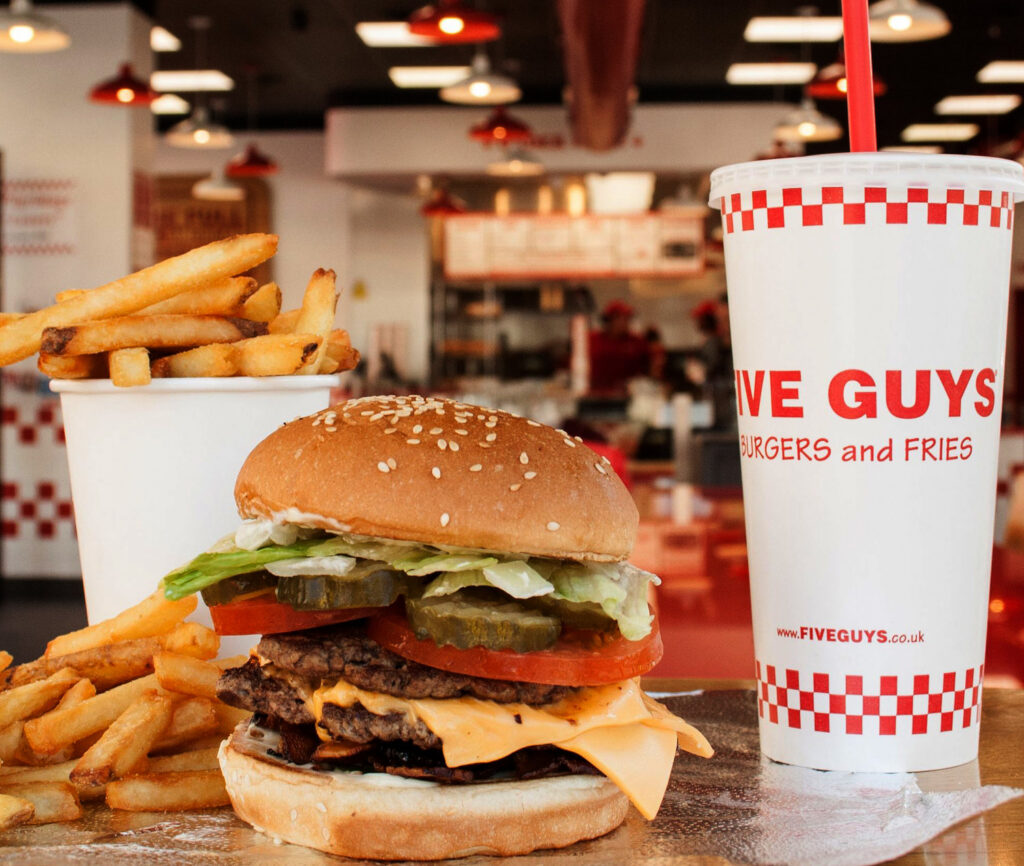 A classic Five Guys meal featuring a double cheeseburger with melted cheese, fresh lettuce, tomato slices, pickles, and a sesame seed bun is placed on a reflective surface. Next to the burger, a large serving of crispy golden fries spills out of a white cup. To the right, a tall Five Guys-branded soft drink cup with a red straw stands prominently. The background showcases the interior of a Five Guys restaurant with its red and white color scheme, bright lighting, and a busy counter area.