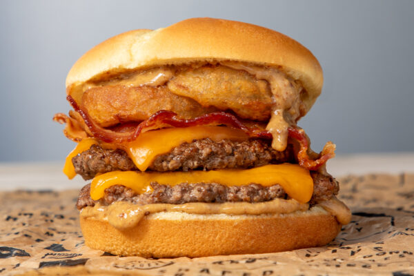 A mouthwatering Wayback Burger stacked high with two beef patties, each topped with melted American cheese. Crispy bacon strips add a smoky crunch, while a golden-fried onion ring sits above the layers, dripping with a creamy, flavorful sauce. The soft, lightly toasted bun holds everything together, with sauce oozing over the edges, making for a visually indulgent and savory burger experience.