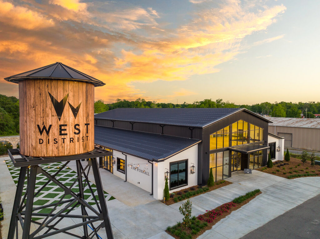 The Foxhall venue located in the West District, featuring a modern industrial-style building with a large glass front entrance. A wooden water tower with 'West District' branding stands prominently to the left. The scene is set against a vibrant sunset sky with lush greenery surrounding the area.
