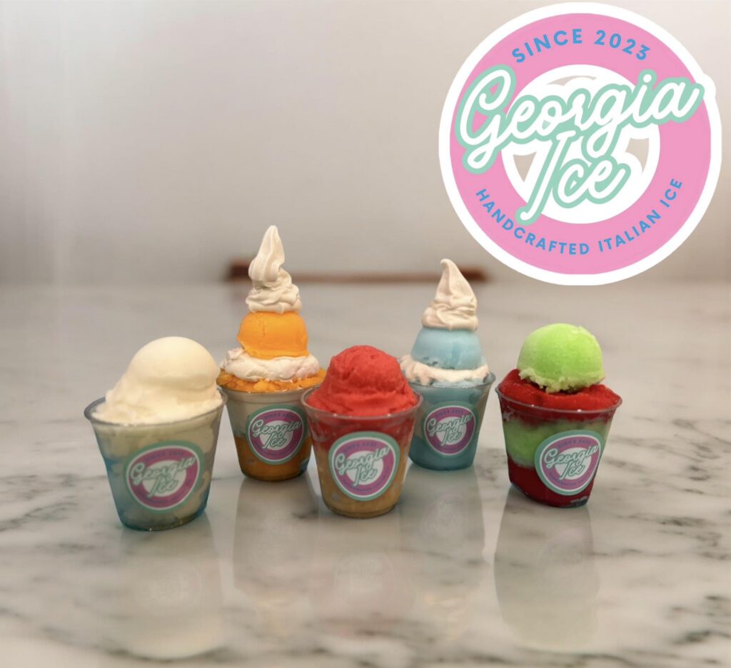 A display of five mini cups of handcrafted Italian ice from Georgia Ice, arranged on a marble countertop. Each cup features a different flavor with vibrant colors, including white, orange, red, blue, and green, some topped with a swirl of whipped cream. The cups have the Georgia Ice logo on the front. In the background, the Georgia Ice logo is prominently displayed with the text 'Since 2023' and 'Handcrafted Italian Ice' in pastel pink and blue colors, reinforcing the brand's identity.