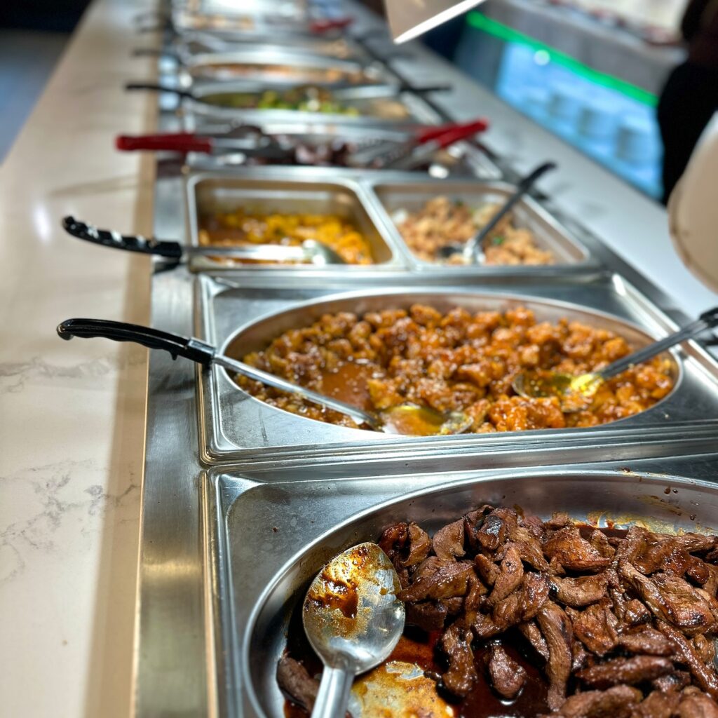 A row of stainless-steel buffet trays filled with Chinese dishes at the Happy Wok Restaurant, featuring glazed meats, stir-fried options, and sauced entrees, each tray equipped with a serving spoon, viewed from an angled perspective along the buffet counter under warm indoor lighting.