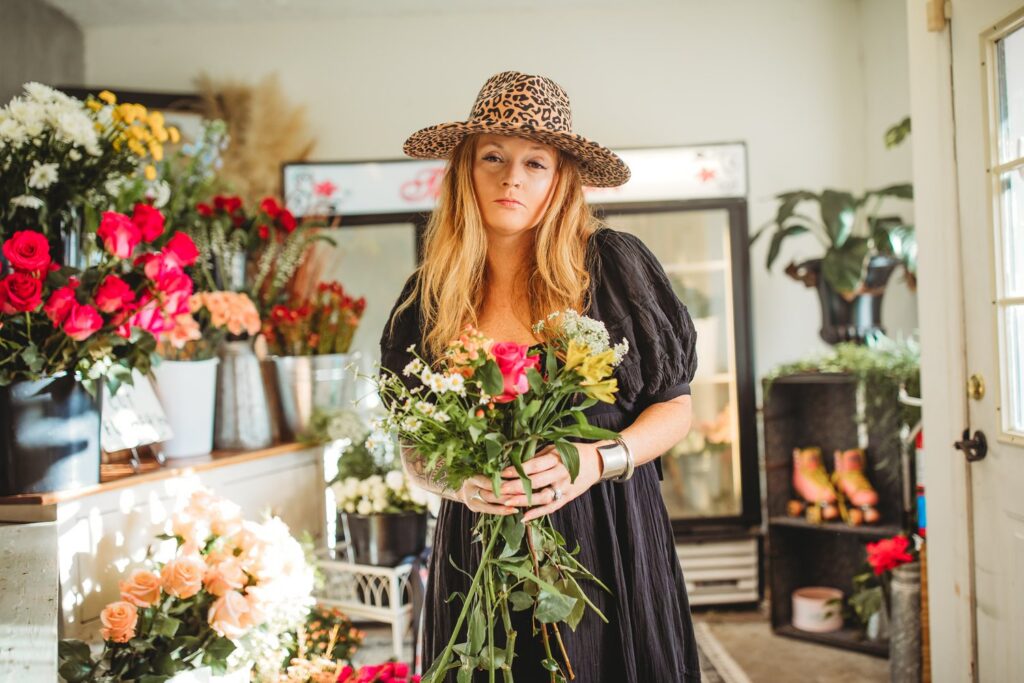 A woman with long, wavy blonde hair stands in a flower shop, holding a bouquet of fresh roses and greenery. She wears a black dress with puffed sleeves, a leopard-print hat, and silver bangles on her wrist. Her expression is calm and confident as she gazes at the camera. The shop is filled with vibrant floral arrangements in buckets and vases, including red, pink, and peach roses. In the background, a refrigerator stocked with flowers, potted plants, and a shelf with vintage roller skates add to the boutique’s charming aesthetic. The Flowergirl has been delivering beauty and memories in downtown Statesboro for nearly 20 years.
