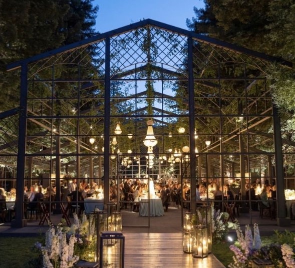 The Atrium on Eighty event venue at dusk, featuring a glass-paneled structure illuminated with warm lights and chandeliers. The interior is filled with guests seated at tables, enjoying an elegant gathering surrounded by lush greenery and decorative floral arrangements.