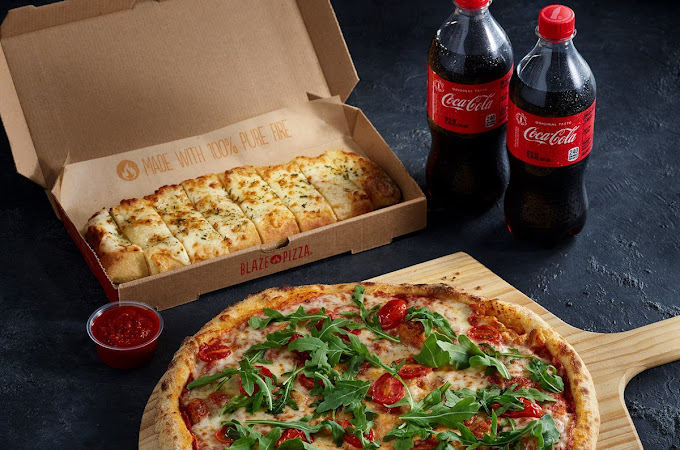 A freshly baked pizza with melted cheese, cherry tomatoes, and arugula is placed on a wooden pizza peel. Beside it, a box of cheesy garlic breadsticks from Blaze Pizza is partially open, revealing golden, seasoned bread slices. A small cup of marinara dipping sauce sits nearby. In the background, two bottles of Coca-Cola are placed on a dark textured surface.