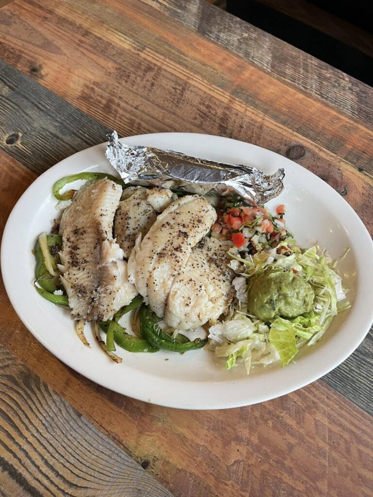 A plated grilled fish dish from Tierra Caliente 1, featuring three seasoned white fish fillets atop sautéed bell peppers and onions. The plate also includes a foil-wrapped tortilla, fresh pico de gallo, shredded lettuce, and a scoop of guacamole, all served on a rustic wooden table. A colorful and fresh Mexican-style seafood option.