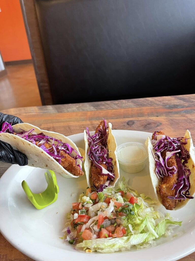A plate of three crispy fish tacos from Tierra Caliente 1, each filled with golden fried fish, shredded red cabbage, and wrapped in soft flour tortillas. A side of shredded lettuce and pico de gallo is served alongside a cup of creamy white sauce. One taco is held by a gloved hand while the others are propped up with a green taco holder on a rustic wooden table, highlighting a fresh and vibrant seafood option.