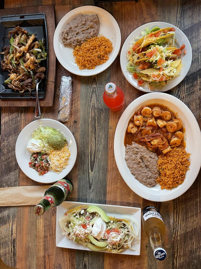 An overhead view of a vibrant Mexican meal spread from Tierra Caliente 1, featuring a sizzling fajita platter with grilled chicken and beef, shrimp in red sauce with rice and beans, crispy tacos topped with shredded lettuce and cheese, a plate of nachos or tostadas with guacamole and sour cream, and traditional sides of Mexican rice, refried beans, and fresh salsa. The table also includes Jarritos soda and bottles of Modelo and Corona beers, all arranged on a rustic wooden surface.