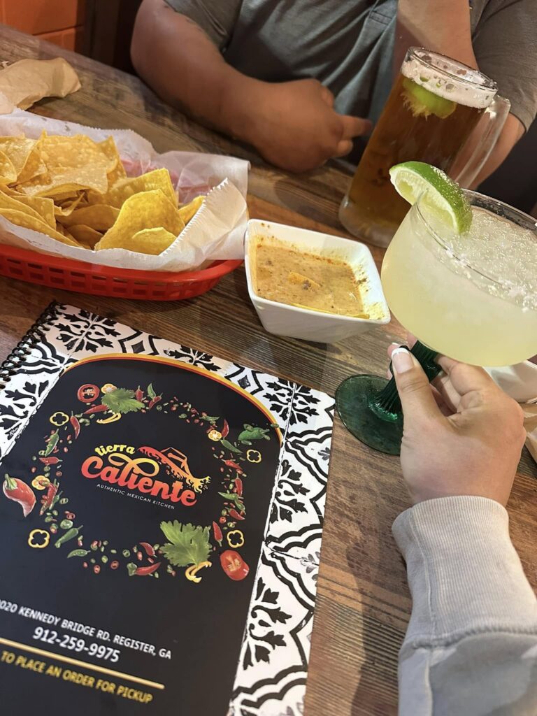 A cozy dining scene at Tierra Caliente 1 featuring a colorful menu, a basket of crispy tortilla chips, a bowl of creamy queso dip, a chilled margarita with a lime wedge, and a glass of iced tea garnished with a lime. The setting captures a relaxed, inviting atmosphere perfect for enjoying authentic Mexican cuisine. The restaurant’s name and contact information are visible on the menu placed on a rustic wooden table.
