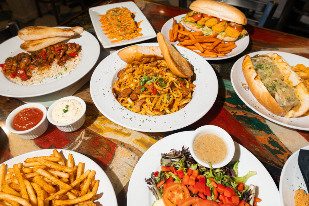 A colorful spread of Southern-style comfort food from Huey’s, featuring a variety of dishes served on a rustic, multi-colored table. The image includes a pasta dish with garlic bread, a loaded po' boy sandwich with fried seafood and sweet potato fries, a Philly cheesesteak, blackened chicken with rice, seasoned fries, a fresh salad with vinaigrette, and two dipping sauces. The presentation highlights Huey’s diverse menu of bold, flavorful offerings.