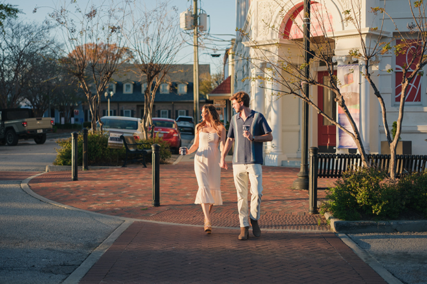 A young couple walks hand in hand through a charming downtown street during golden hour, each holding a drink cup. The woman wears a light, elegant dress and heels, while the man is dressed casually in a short-sleeve button-down and pants. They smile and enjoy the relaxed evening atmosphere. The background features a historic building with red and white accents, brick sidewalks, and quaint storefronts—capturing the essence of the Sip and Soar event, which celebrates community, shopping, and local flavor in a lively, walkable setting.