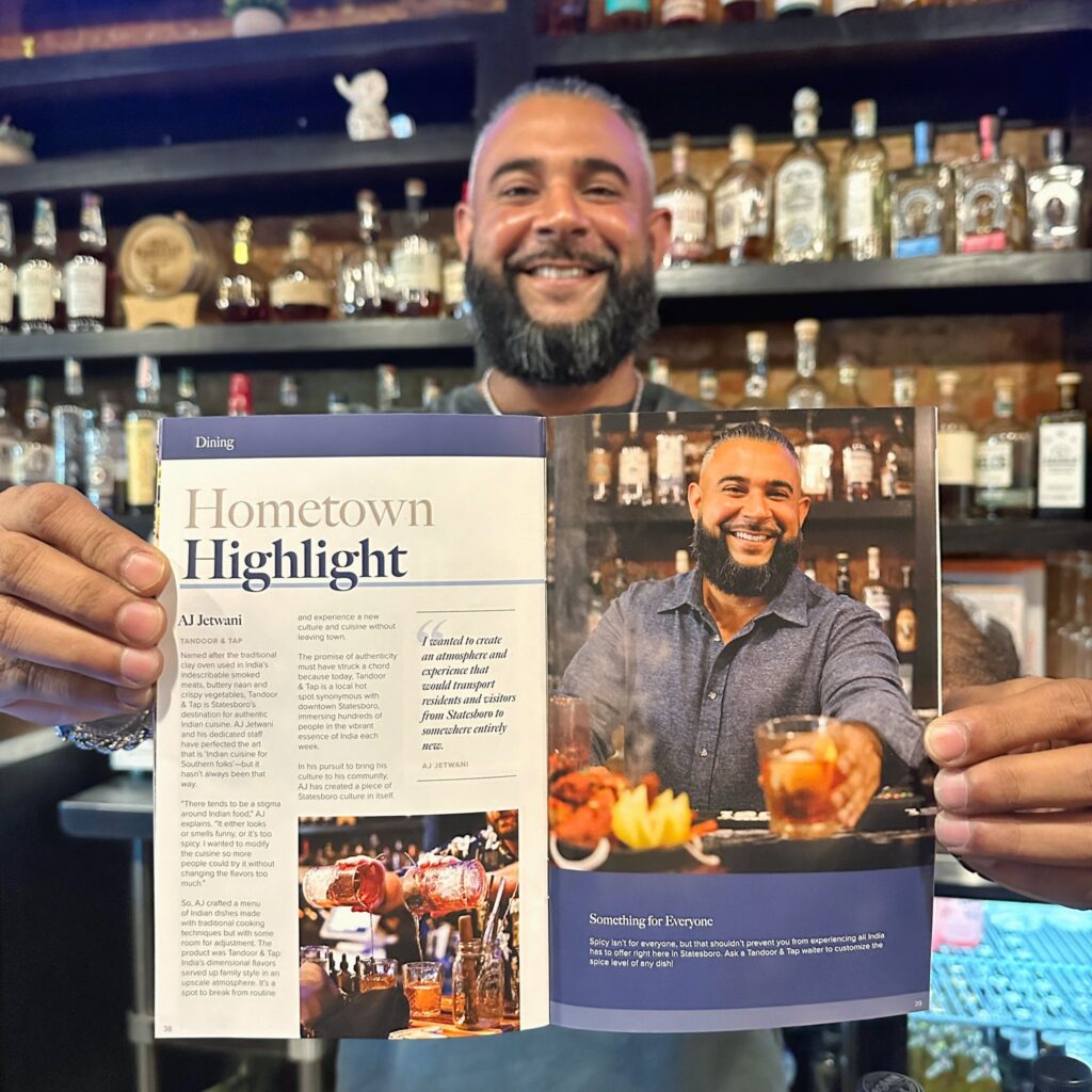 A smiling man proudly holds open the Statesboro Visitors Guide, displaying a feature titled “Hometown Highlight” about AJ Jetwani, who stands behind the magazine in the same pose, creating a mirrored moment. The article highlights Jetwani’s impact on the local dining scene, with a quote about creating a welcoming atmosphere that reflects Statesboro’s culture. He stands in front of a shelf full of liquor bottles, reinforcing his role in the hospitality industry. This image is part of the celebration for Visit Statesboro winning the prestigious Communicator Award for the 2024 Visitors Guide.