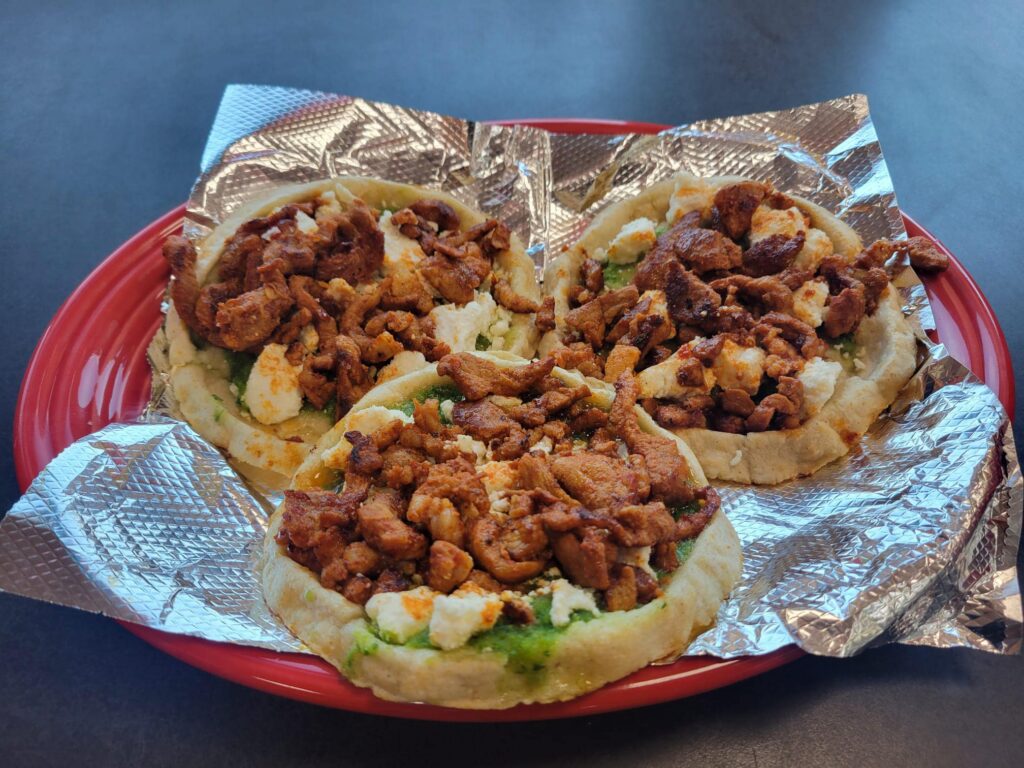 A red plate holds three freshly made Mexican sopes from Zerrano’s Taqueria, each topped with a layer of green salsa, crumbled cheese, and a generous portion of seasoned grilled meat. The sopes rest on pieces of foil and sit atop a dark table surface.
