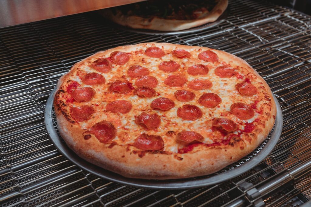 A freshly baked pepperoni pizza from Holy Pie! Pizzeria is shown on a metal tray inside an oven. The pizza features a golden, crispy crust with melted mozzarella cheese and evenly spaced slices of pepperoni. Another pizza can be seen baking on a rack above in the background.