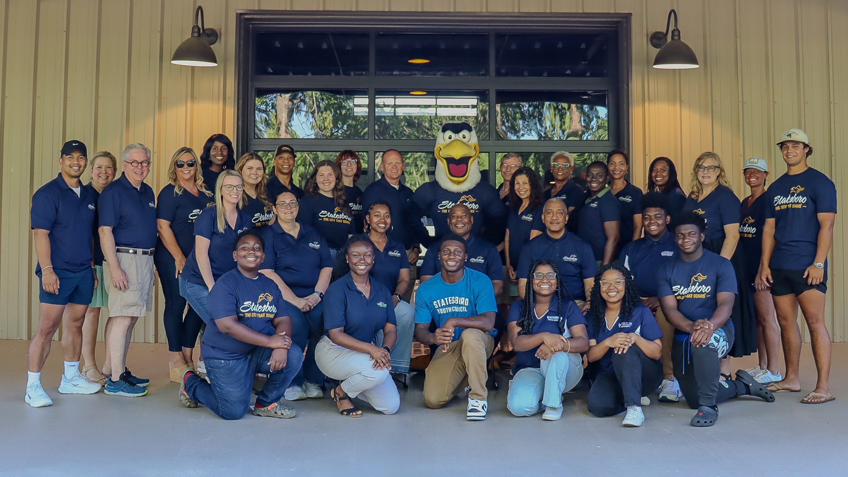 A diverse group of adults pose together in front of a building with a large window, all wearing matching navy blue shirts that read “Statesboro: The City That Soars.” The group includes men, women, and young adults, smiling and kneeling or standing in rows. A cheerful eagle mascot is in the center of the back row. The group appears to be part of a community or civic organization, possibly preparing to participate in the All-America City Competition.