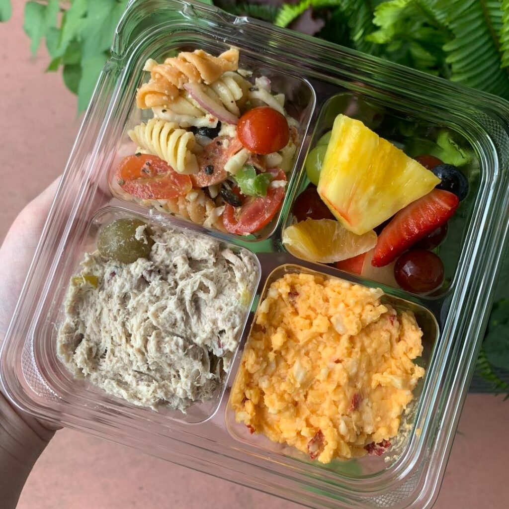 Sampler tray from All the Fixin’s: clear clamshell with four sections—rotini pasta salad with cherry tomatoes, mixed fruit (pineapple, strawberries, grapes, orange, blueberry), a creamy deli salad topped with an olive, and a pimento-cheese style spread.