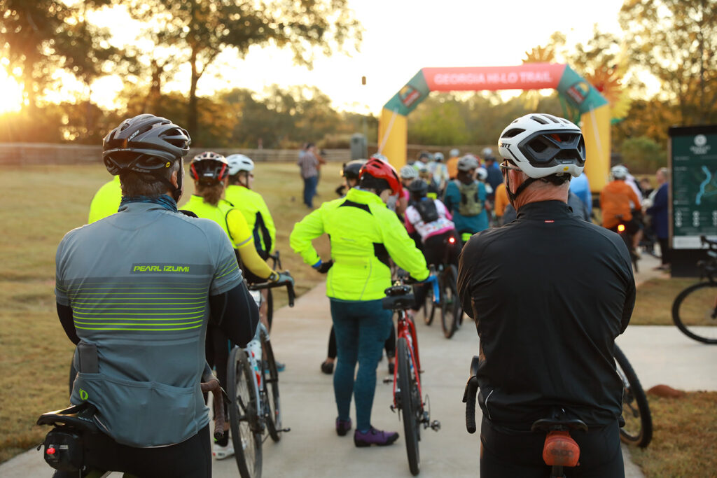 Cyclists gather with their bikes at the starting area of the Georgia Hi-Lo Trail Transitions ride, wearing helmets and bright cycling gear as they line up beneath a Georgia Hi-Lo Trail arch at sunrise, capturing the anticipation, community, and early-morning atmosphere of the Athens to Savannah cycling event.