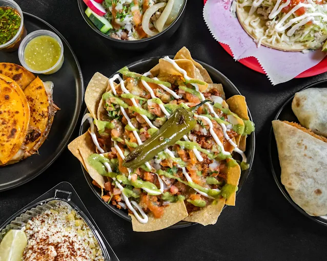 An overhead photo of a spread of Mexican-inspired dishes from Flacos, featuring a bowl of loaded nachos topped with guacamole, sour cream, pico de gallo, and a whole jalapeño, surrounded by tacos, burritos, quesadillas, and small containers of salsa and green sauce on a dark tabletop.