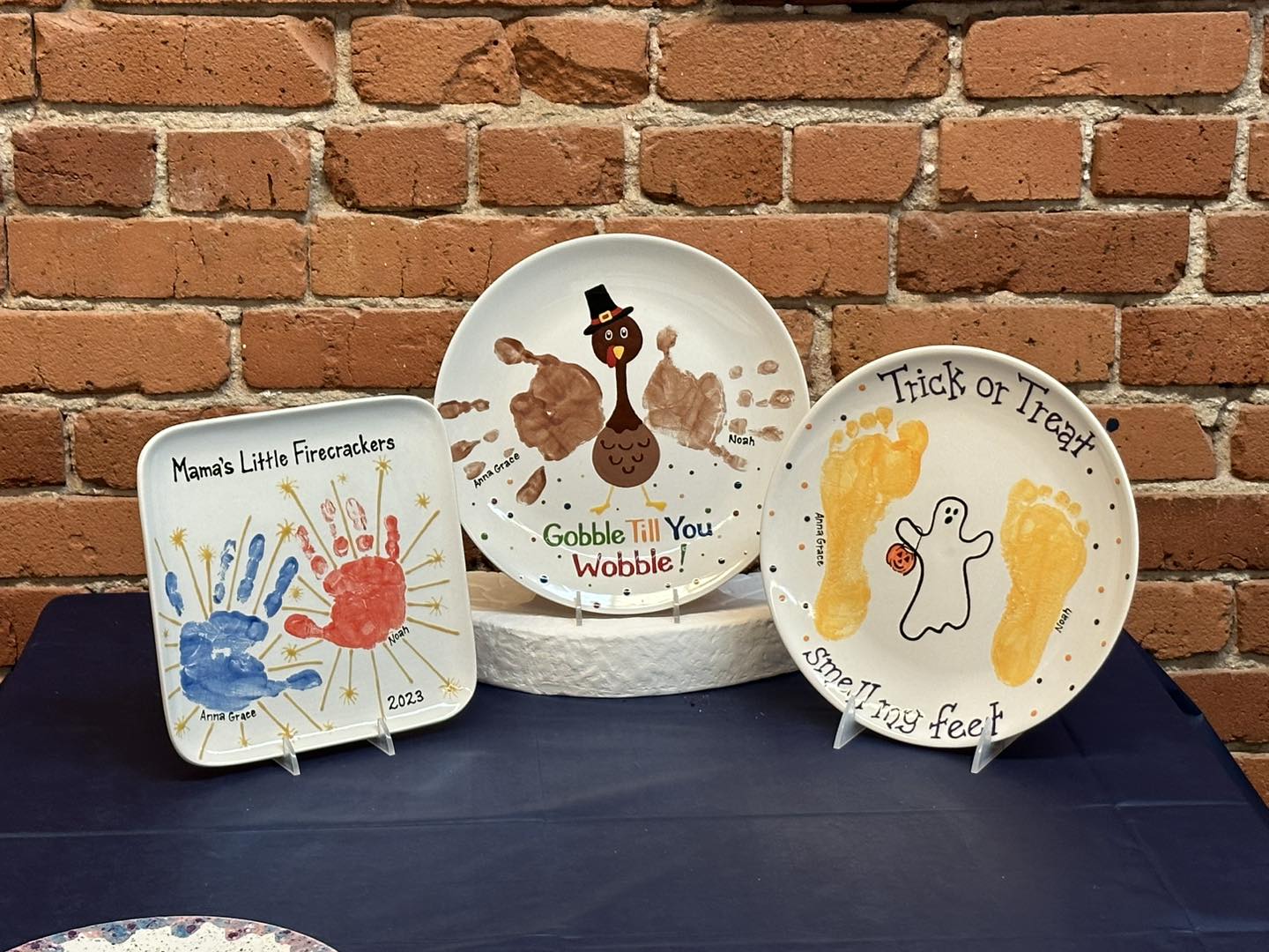 Three decorative plates are displayed on stands against a red brick wall, each featuring handprint or footprint art for different holidays. The left square plate reads “Mama’s Little Firecrackers” and has red and blue handprints with yellow spark designs labeled with the names Anna Grace and Noah and the year 2023. The center round plate shows a Thanksgiving turkey made from brown handprints with the words “Gobble Till You Wobble!” written below. The right round plate has two yellow footprints forming a Halloween design with a white ghost between them, labeled “Trick or Treat Smelly Feet.” All three sit on a dark blue tablecloth, creating a festive and colorful display.