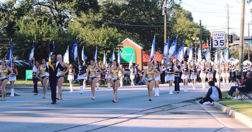 A lively parade scene on a sunny day shows a large marching band (The Georgia southern marching band) and dance line performing down a city street. The front row features uniformed dancers in gold and white costumes leading with synchronized high-step movements beside a drum major in a navy and gold outfit. Behind them, more performers in matching attire carry blue and white flags while the band follows farther back playing instruments. Spectators sit and watch along the sidewalks as trees and brick buildings line the background under a clear blue sky.