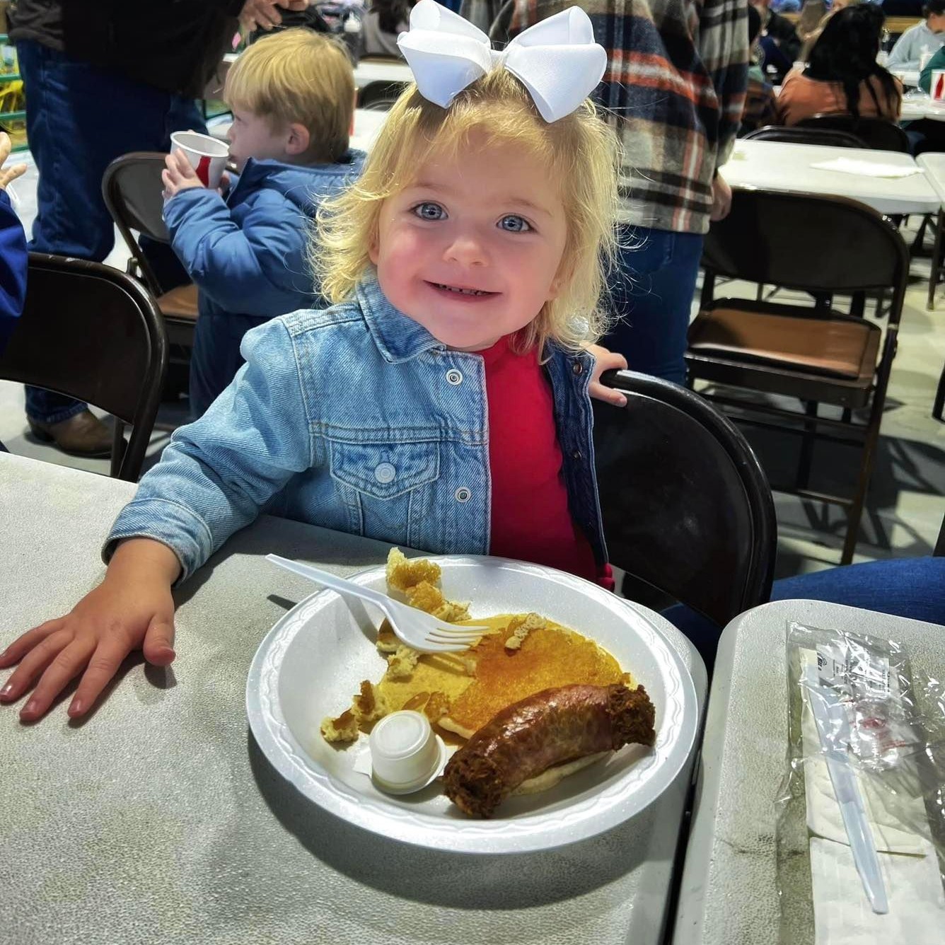 A young girl with blonde hair, a large white bow, and a denim jacket over a red shirt smiles while sitting at a table during the Kiwanis Ogeechee Fair. In front of her is a paper plate with pancakes, a sausage link, a small syrup cup, and a plastic fork. She sits in a crowded indoor area filled with people and folding chairs, capturing a cheerful moment at the community breakfast event.