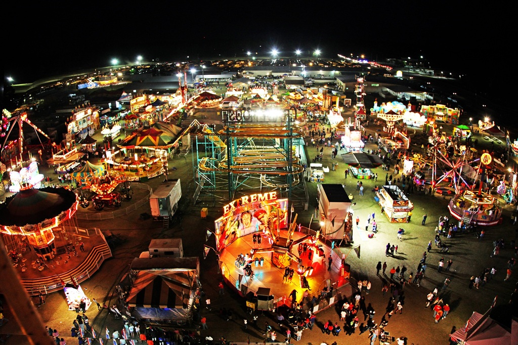 A vibrant nighttime aerial view of the Kiwanis Ogeechee Fair shows a sprawling fairground filled with colorful lights, rides, and game booths. Brightly lit attractions like carousels, roller coasters, and spinning rides glow against the dark sky while crowds of people walk along the pathways enjoying the festivities. Food stands and tents are scattered throughout the fair, creating a lively and energetic atmosphere typical of a bustling community fair.