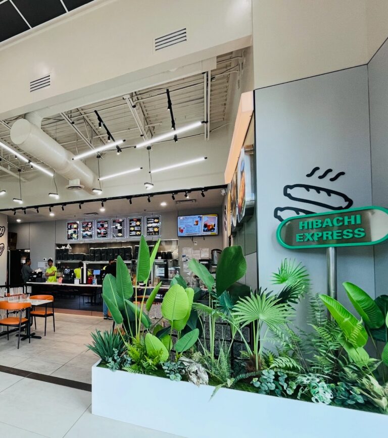An interior photo of Hibachi Express Ramen showing the restaurant’s counter and ordering area, with illuminated menu boards, seating in the background, and a green “Hibachi Express” wall sign beside a planter filled with lush indoor greenery, creating a modern and inviting dining atmosphere.