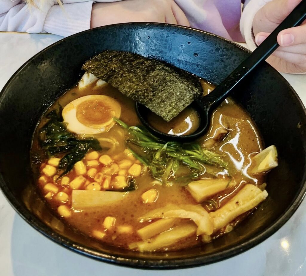 A close-up photo of a bowl of ramen from Hibachi Express Ramen, featuring a rich broth with noodles, a halved soft-boiled egg, corn, bamboo shoots, leafy greens, and a sheet of seaweed, with a black soup spoon resting inside the bowl.