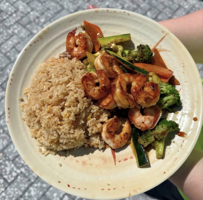 A close-up food photo from Hibachi Express Ramen showing a plate of hibachi-style shrimp served with fried rice and a mix of sautéed vegetables, including broccoli, zucchini, and carrots, lightly glazed with sauce and held outdoors in natural sunlight.