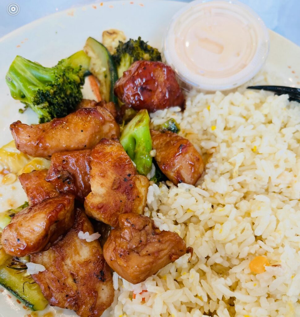 A close-up food photo from Hibachi Express Ramen featuring hibachi-style grilled chicken served with white rice and sautéed vegetables, including broccoli and zucchini, accompanied by a small cup of sauce on a white plate under bright lighting.