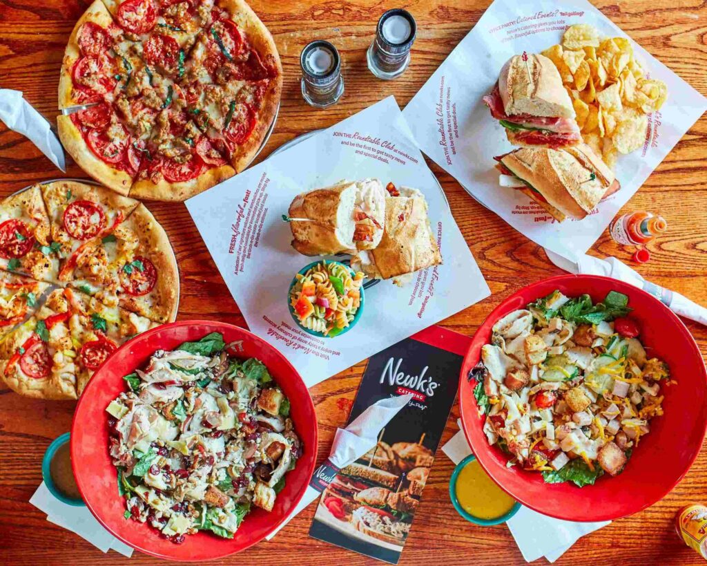 An overhead promotional photo showcasing a variety of menu items from Newk’s Eatery, including artisan pizzas, fresh salads in red bowls, sliced sandwiches with chips, and a pasta dish, all arranged on a wooden table with menus, seasonings, and drinks, highlighting the restaurant’s fresh and casual dining options.