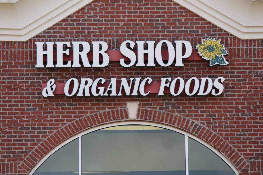 The Herb Shop & Organic Foods storefront sign is mounted on a red brick building above an arched window. The large white lettering sits on red panels and is accompanied by a yellow flower logo, highlighting the store’s focus on natural herbs, organic foods, and wellness products.