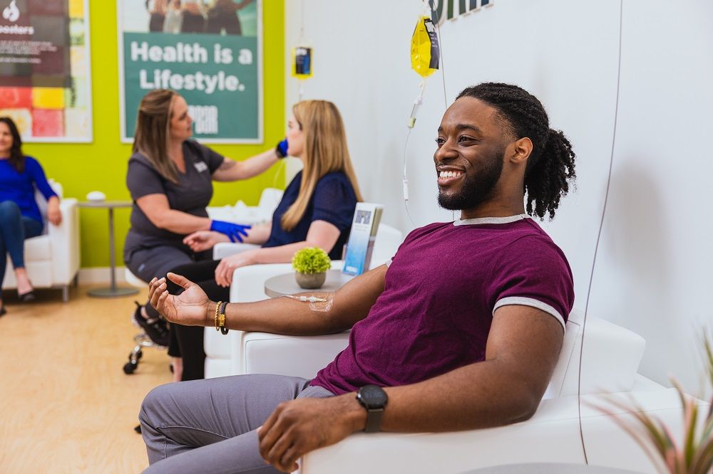 A man sits comfortably in a white lounge chair at The Drip Bar while receiving an IV vitamin infusion, smiling as the drip bag hangs beside him. In the background, a staff member wearing gloves assists another client with a treatment. The modern wellness clinic features bright green walls, clean décor, and a sign reading “Health Is a Lifestyle,” creating a relaxed and professional health-focused atmosphere.