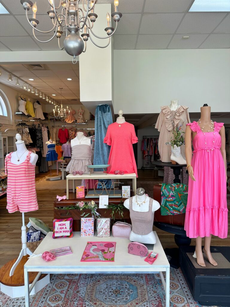 Inside The Collective & Vine clothing store, mannequins display colorful dresses and summer outfits while tables showcase jewelry, handbags, and accessories. The boutique features warm wooden floors, elegant lighting, and neatly arranged racks of clothing throughout the bright and stylish retail space.