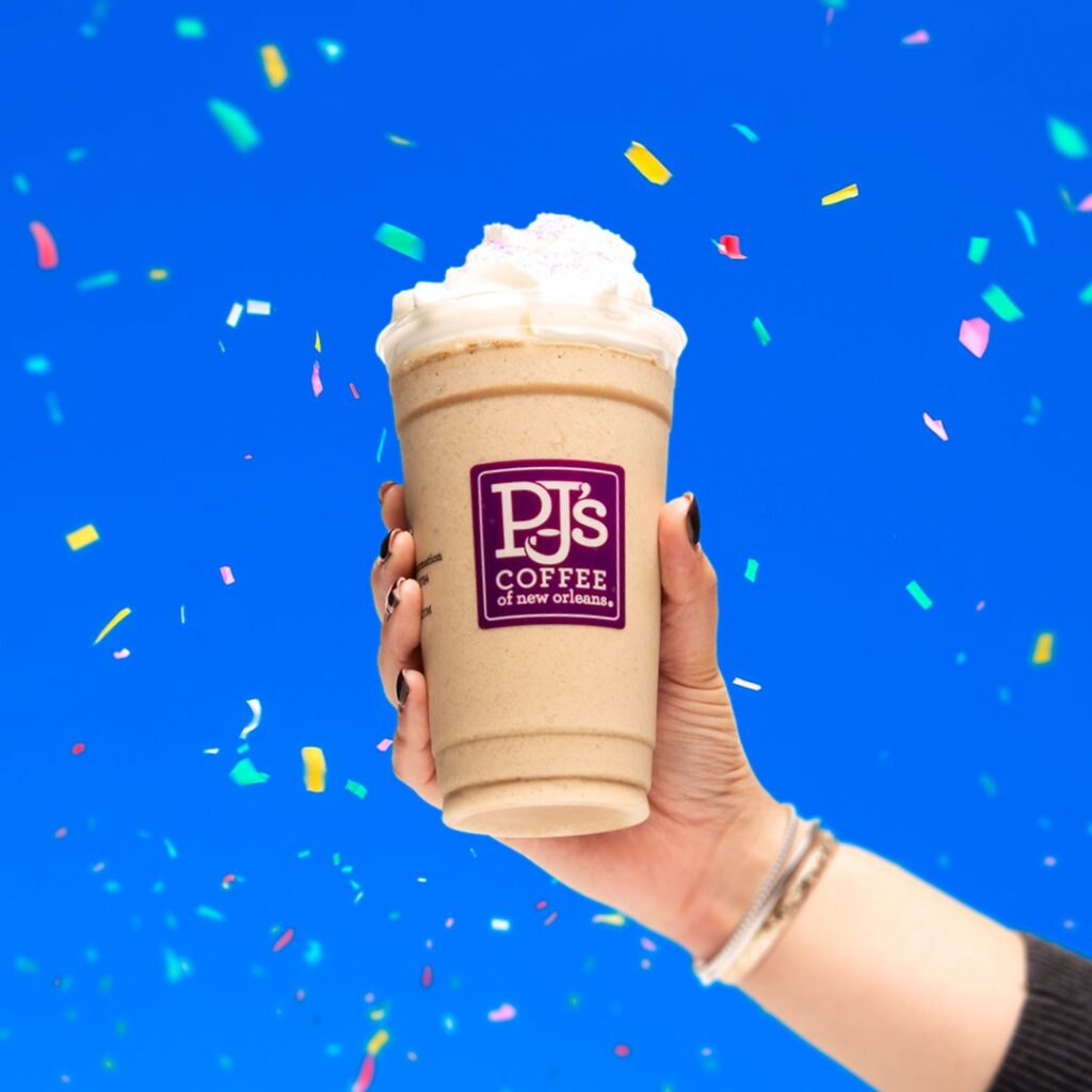 Frozen blended coffee drink from PJ’s Coffee topped with whipped cream and held in front of a bright blue background with colorful confetti.